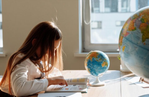 Ett barn med långt hår läser i en bok vid ett bord där det står två jordglober.